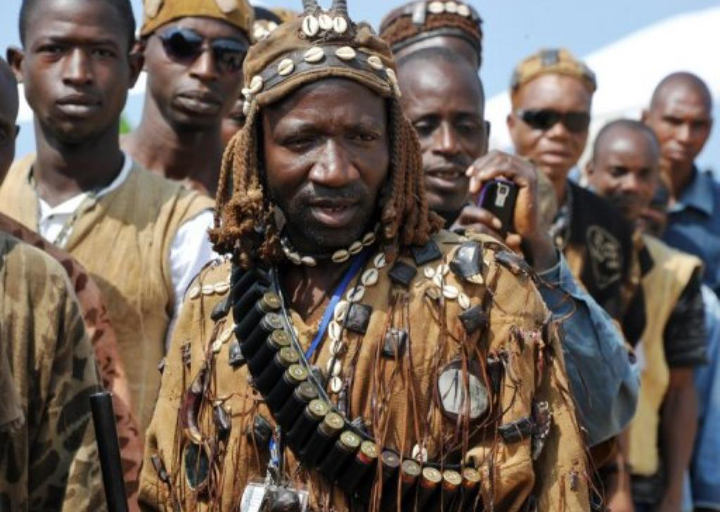 Several men in traditional Malian clothes. The man in the front is wearing a hunter tunic decorated with cowrie shells and tassels, and is wearing a brown hat with a leather strip with cowrie shells attached.