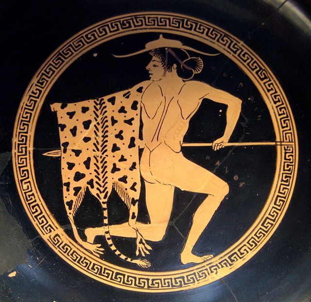 An image of a man wearing a wide brimmed petasos and a leopard skin over his shoulder. The man is drawn in light orange, and there's a circular border with a Greek pattern as well. The background is black.